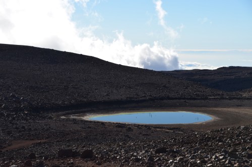 A serene, reflective pond surrounded by rocky terrain under a clear blue sky with scattered clouds.