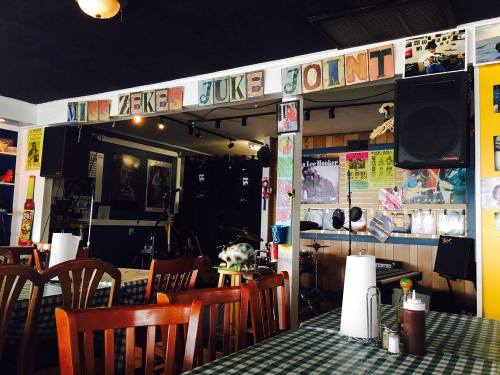 Interior of a juke joint with wooden tables, checkered tablecloths, and a stage for live music in the background.