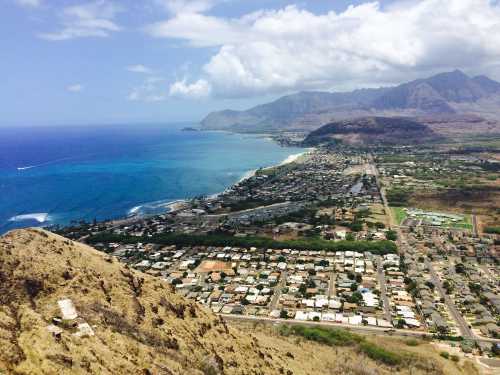 Aerial view of a coastal town with blue ocean, sandy beach, and mountains under a partly cloudy sky.