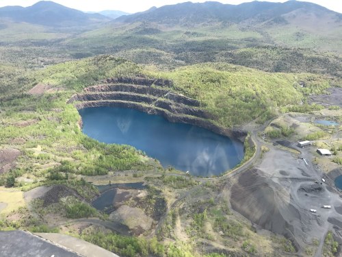 Aerial view of a deep blue lake surrounded by green hills and rocky terrain, with mining areas visible nearby.