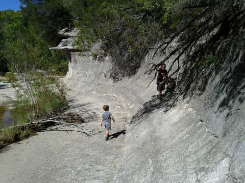 Two children explore a rocky path by a river, surrounded by trees and greenery on a sunny day.