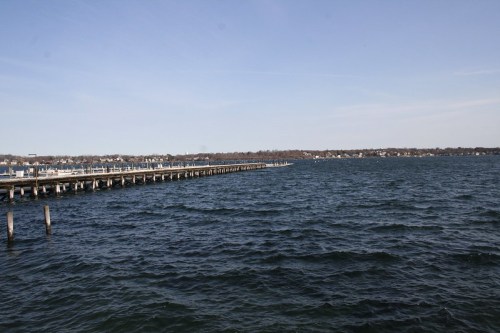A wooden pier extends into a calm, blue lake under a clear sky, with distant shoreline visible.