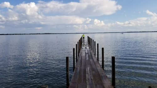 A person walks along a wooden pier extending over calm water under a cloudy sky.
