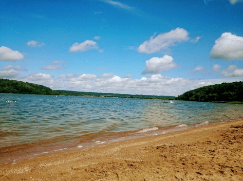 A serene beach scene with sandy shore, calm water, and a blue sky dotted with fluffy clouds. Green hills in the background.