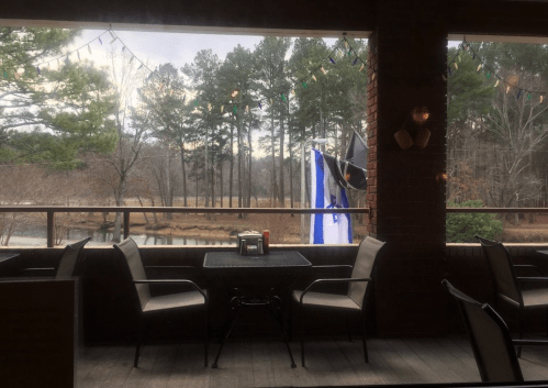 View from a porch with empty chairs, overlooking a wooded area and a calm body of water, with string lights in the background.
