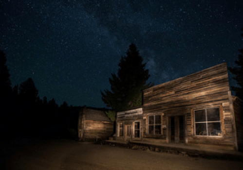 A rustic wooden building under a starry night sky, surrounded by trees in a quiet, dark setting.