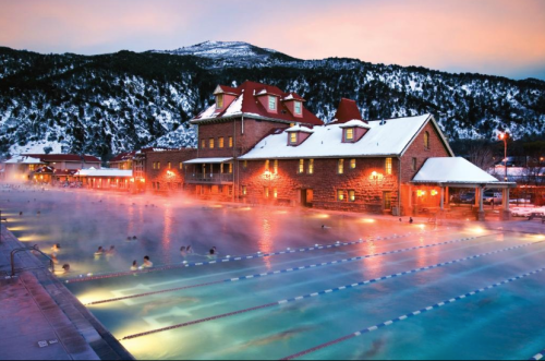 A scenic outdoor pool surrounded by mountains, with steam rising and a historic building illuminated at sunset.