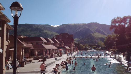 A sunny pool scene with people swimming, surrounded by mountains and buildings, creating a relaxing atmosphere.