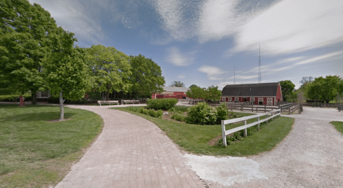 A scenic view of a farm with red barns, green trees, and a winding brick path under a blue sky.