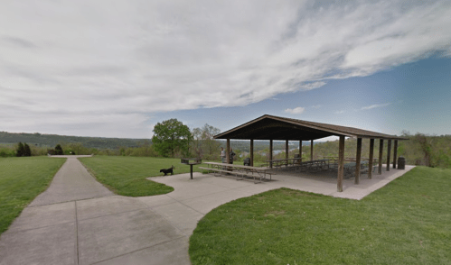 A grassy area with a covered picnic shelter and a path, overlooking a scenic landscape under a cloudy sky.