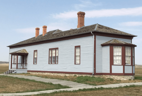 A historic blue wooden house with a porch and multiple chimneys, set against a clear sky and grassy landscape.