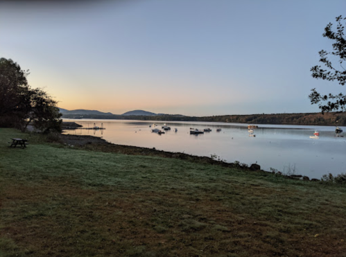 Serene lakeside view at dawn, with boats anchored and mountains in the background, surrounded by lush greenery.