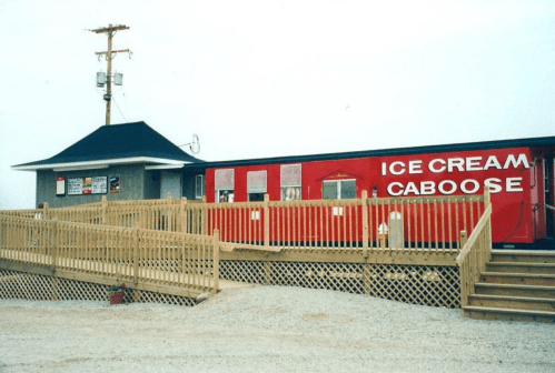 A red ice cream caboose with a wooden deck, featuring signs and a small building beside it.