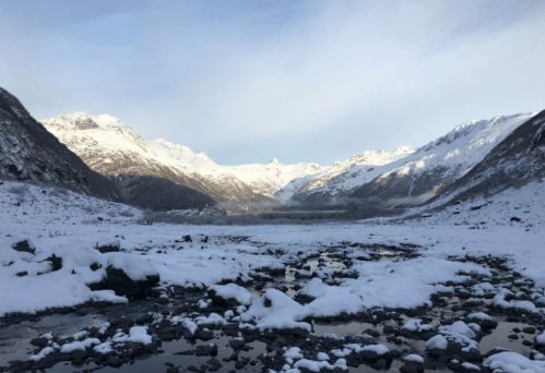 Snow-covered mountains surround a rocky landscape, with a calm river reflecting the serene winter scenery.