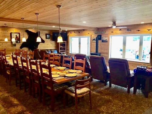 A spacious dining area with a long table, wooden chairs, and large windows, featuring rustic decor and a bear mount.