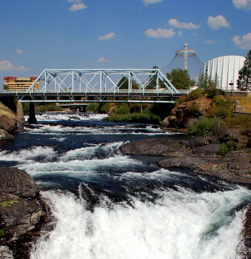 A blue bridge spans a rushing river with rocky banks, surrounded by greenery and buildings under a blue sky.
