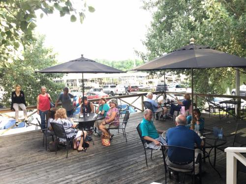 A lively outdoor dining area with people sitting under umbrellas, surrounded by trees and a view of parked boats.