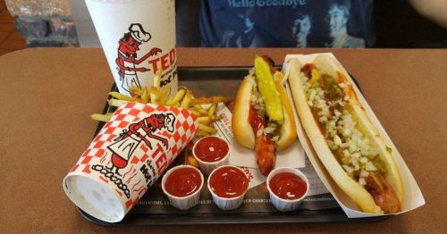 A tray with two hot dogs topped with onions and pickles, fries, and a drink, all from Ted's.