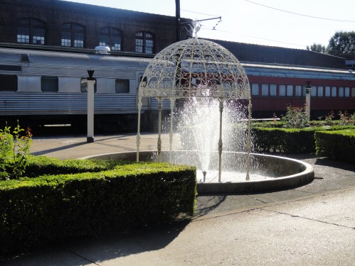 A decorative white fountain surrounded by hedges, with a vintage train in the background under bright sunlight.
