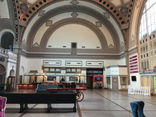 Interior of a spacious hall with high ceilings, large windows, and a reception area featuring a vintage cart.