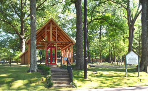 A wooden pavilion with red doors surrounded by trees and greenery, featuring a sign nearby.