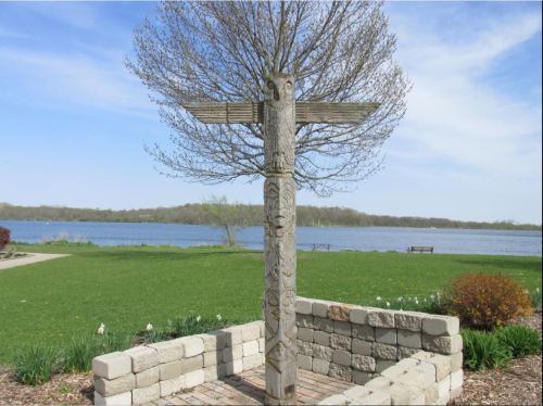 A carved totem pole stands in a park by a lake, surrounded by greenery and flowers under a clear blue sky.