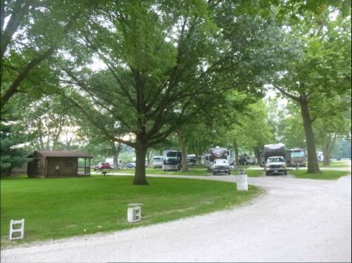 A gravel road winds through a green campground with RVs parked under large trees. A small cabin is visible nearby.