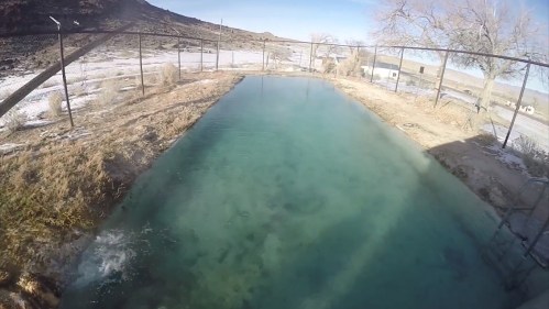 A clear, turquoise pool surrounded by dry land and sparse vegetation under a bright blue sky.