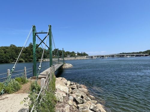 A green suspension bridge spans a calm river, with rocky banks and trees in the background under a clear blue sky.