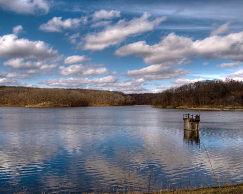 A serene lake surrounded by trees, with a cloudy blue sky reflecting on the water's surface.
