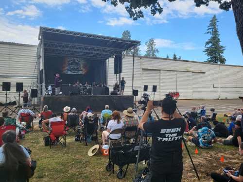 A crowd watches a live performance on an outdoor stage, with a camera operator in the foreground.