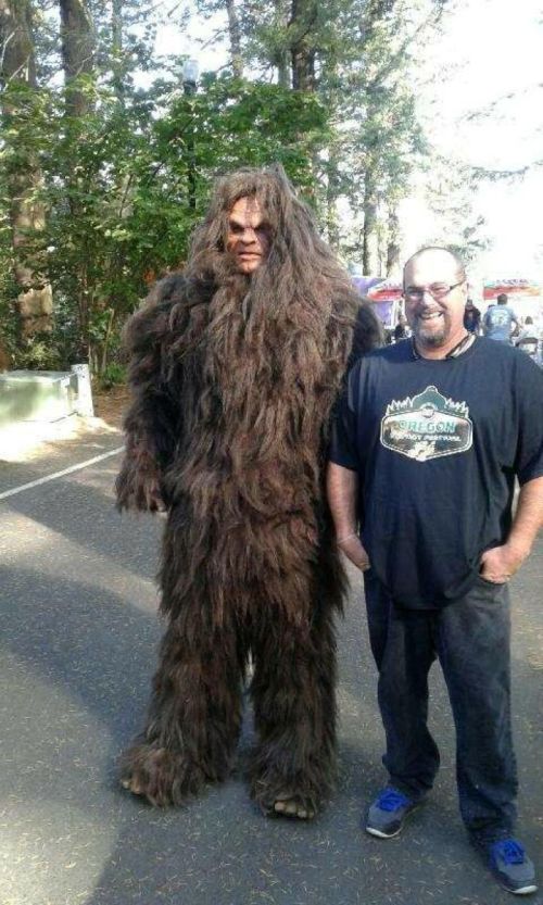 A person in a Bigfoot costume stands next to a smiling man in a black t-shirt, surrounded by trees.