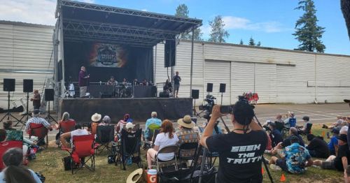 A crowd watches a live band perform on stage at an outdoor event, with a person filming from the front.