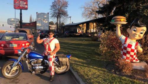 A man in a checkered outfit poses on a motorcycle next to a large burger mascot outside a restaurant.