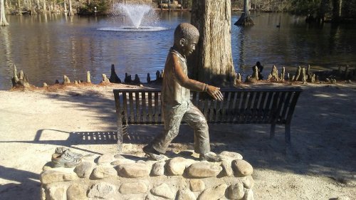 A bronze statue of a boy walking beside a pond, with a bench and fountain in the background.