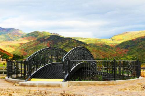 A decorative black bridge arches over a scenic landscape of rolling hills and colorful foliage under a cloudy sky.