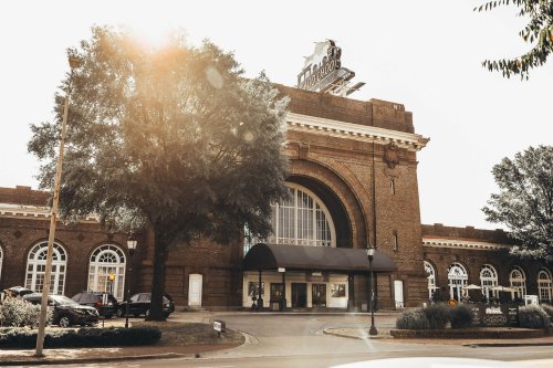 Historic train station with a large archway entrance, surrounded by trees and parked cars, under a sunny sky.