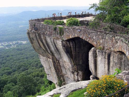A scenic overlook with a stone railing, surrounded by greenery and mountains in the background.