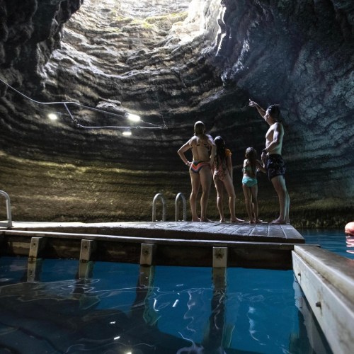 A group of people stands on a wooden dock inside a cavern, with a pool of blue water and a high, rocky ceiling above.