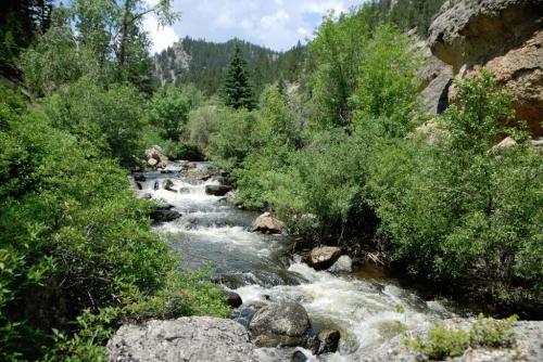 A serene river flows through a lush green landscape, surrounded by trees and rocky terrain under a partly cloudy sky.