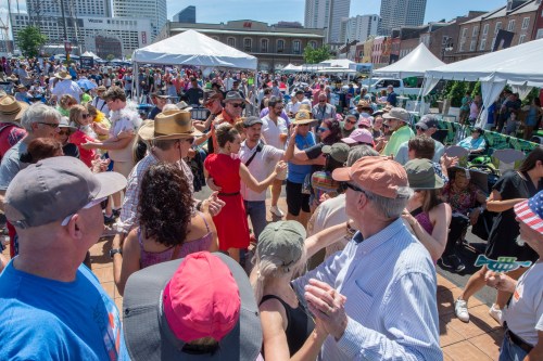 A lively crowd dances at an outdoor festival, with tents and city buildings in the background under a clear blue sky.