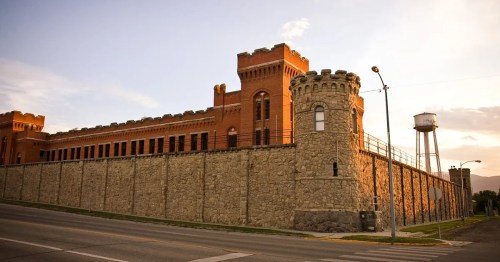 Historic brick building with towers, surrounded by a stone wall, near a water tower and street at sunset.