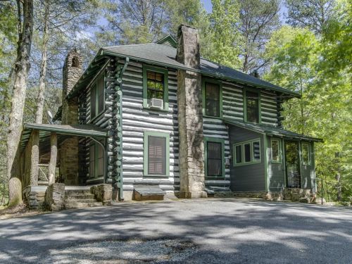 A rustic log cabin surrounded by trees, featuring a stone chimney and a spacious porch.