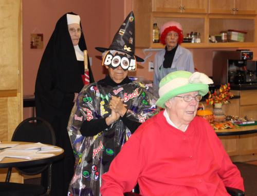 A group of elderly women in costumes, including a witch and a nun, celebrating with festive decorations in a community room.