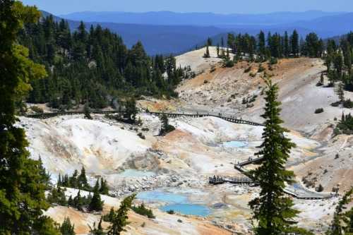 A colorful geothermal landscape with pools and wooden walkways, surrounded by trees and distant mountains.