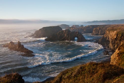 Coastal landscape with rocky formations, waves crashing, and a misty horizon under a clear sky.