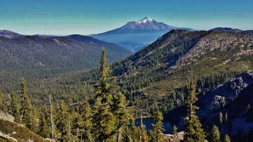 A scenic mountain landscape featuring a snow-capped peak, lush green forests, and a serene lake in the valley below.