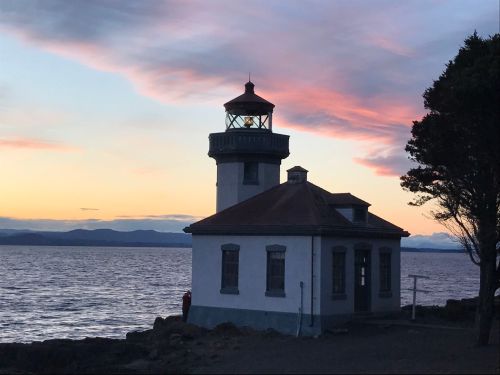 A lighthouse stands by the water at sunset, with colorful clouds and mountains in the background.