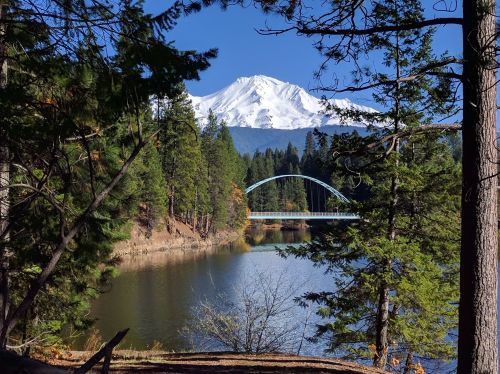 A serene landscape featuring a lake, a bridge, and a snow-capped mountain, framed by lush green trees.
