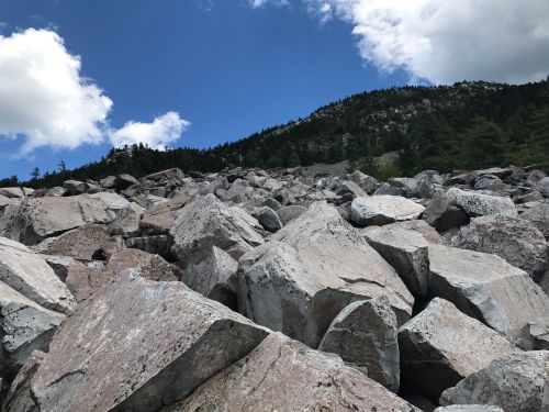A rocky landscape with large boulders under a blue sky and scattered clouds, surrounded by green trees and a mountain.